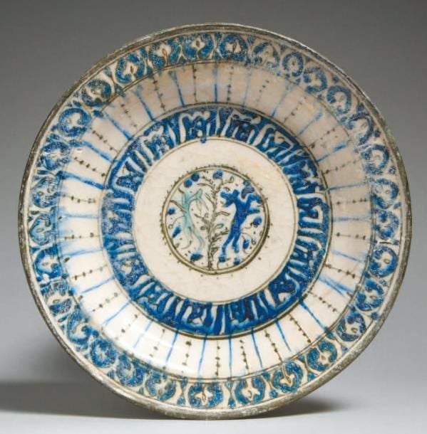 ASSIETTE AUX LIÈVRES Belle assiette blanche, décorée en bleu et turquoise de deux lièvres dans un rondeau et d'un bandeau concentrique d'inscriptions cursives en réserve sur fond bleu, de lignes rayonnantes bleues ou ponctuées noires le long du cavet et d'une frise de palmettes en bordur