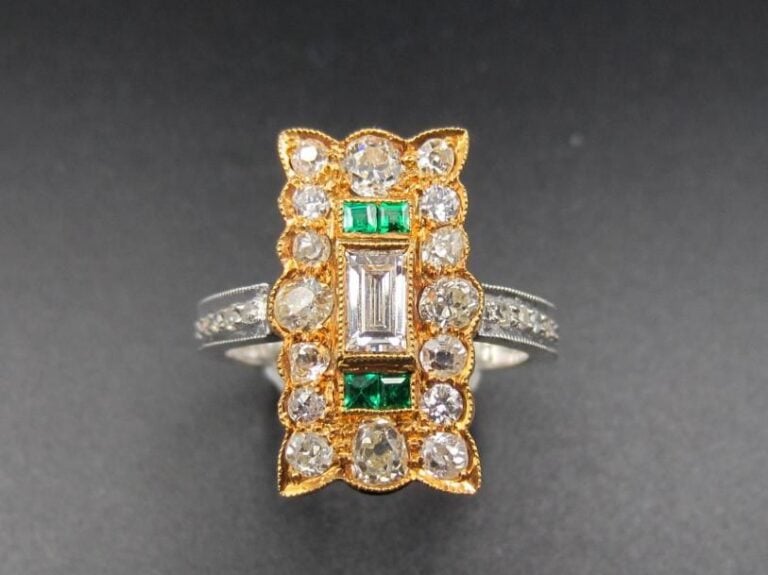 BAGUE en gris et or jaune (750 millièmes) serti d’un diamant baguette central épaulé de quatre émeraudes calibrées, entourées de diamants             Doigt : 53        Poids brut : 6 g