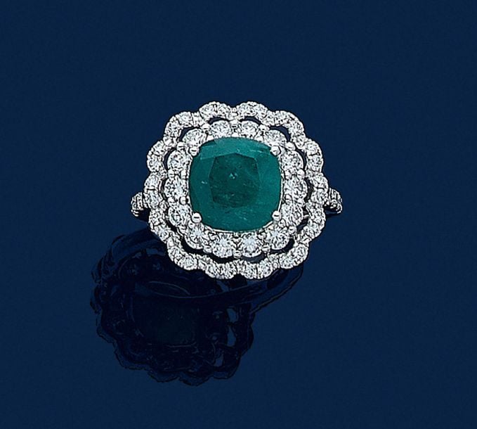 •BAGUE «fleur» en or gris (750‰) ajouré, serti d'une émeraude de forme coussin dans un double entourage de diamants taille brillan