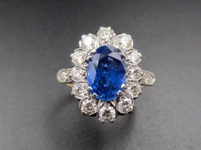 BAGUE « marguerite » en or jaune et or gris (750 millièmes) serti d’un saphir de forme ovale pesant 3,18 carats, entouré et épaulé de diamants taille brillant             Le saphir est accompagné d’un rapport d’analyse gemmologique du laboratoire G       G       T       , n°8020676, datant du 29      2      2016, attestant son poids de 3,18 carats, sa couleur naturelle « Blue», chauffé        Origine : Sri Lanka             Doigt : 58        Poids brut : 9,8 g