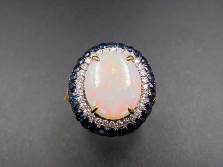 BAGUE ovale en or gris et or noirci (750 millièmes) serti d’une opale entourée de diamants taille brillant et saphirs             Doigt : 55         Poids brut : 4,6 g