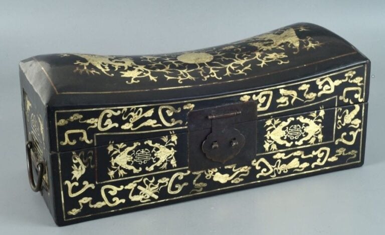 BOITE A BIJOUX EN BOIS LAQUE ET DORE EN FORME D'OREILLER Chine De section rectangulaire, le couvercle incurvé à l'imitation des oreillers Chinois, le devant orné des symboles des huit immortels secrets et de deux chauve-souris affrontées de part et d'autre d'un caractère shou (rébus "puissiez-vous connaître bonheur et longévité"), les côtés de même, le couvercle centré d'une pêche de longévité qu'encadrent une chauve-souris et deux phénix affrontés, pourvue de poignées latérales pendantes (petits accidents, dorure estompée par endroits