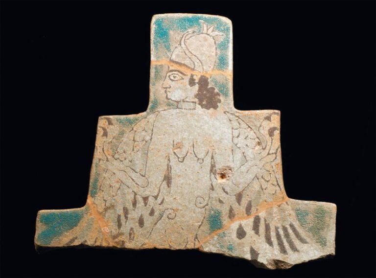 BRIQUE Brique à profil pyramidal étagé, décorée sur fond turquoise d'un personnage ailé, torse nu, la tête de profil coiffée d'un casque conique cornu, sur des cheveux bouclés tombant sur la nuque, paré d'un collier, tenant bras écartés deux « fleurs
