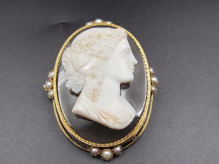 BROCHE ovale en or jaune (750 millièmes) ciselé, serti d’un camée en calcédoine grise deux couches « profil de femme » souligné d’émail noir (usures et restauration) et ponctué de douze demi-perle
