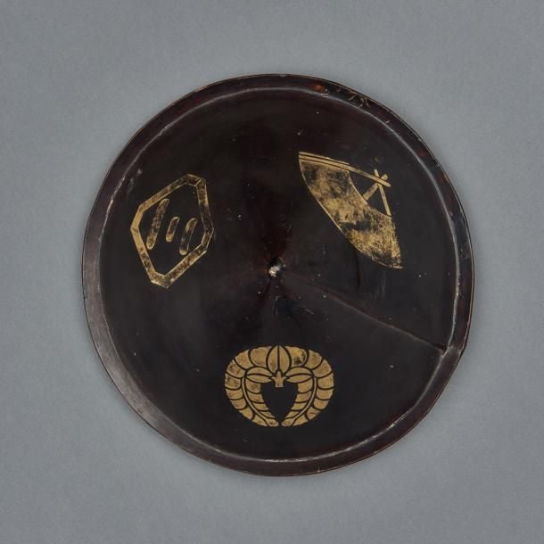 Casque de guerrier japonais (jingasa) de forme conique et de couleur noire, décoré de trois emblèmes en laque maki-e dorée: Un éventail, un wistéria (sagarifuji) ainsi que le caractère «trois» dans un losange aux bords coupés (sumikirikaku ni san) et une bordure vertical