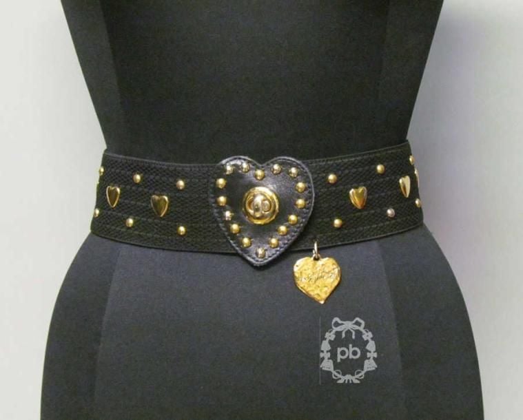 CEINTURE élastique noire ornée de clous et de coeurs en métal doré, fermoir pivotant figurant un coeur en cuir, breloque siglée (T2)