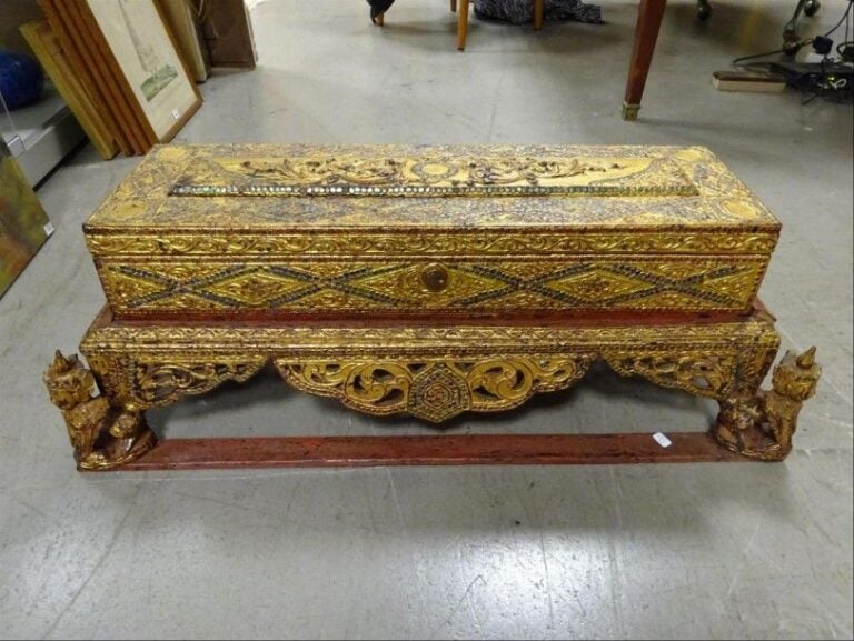 COFFRE / BOITE / RELIQUAIRE DE SECTION RECTANGULAIRE De style, Sud-est asiatique En bois doré richement sculpté aux multiples motifs géométriques et à incrustations, le dessus du couvercle aux losanges inscrustés de pierres colorées, repose sur un socle en bois aux tabliers et ajourés et aux pieds formant déité