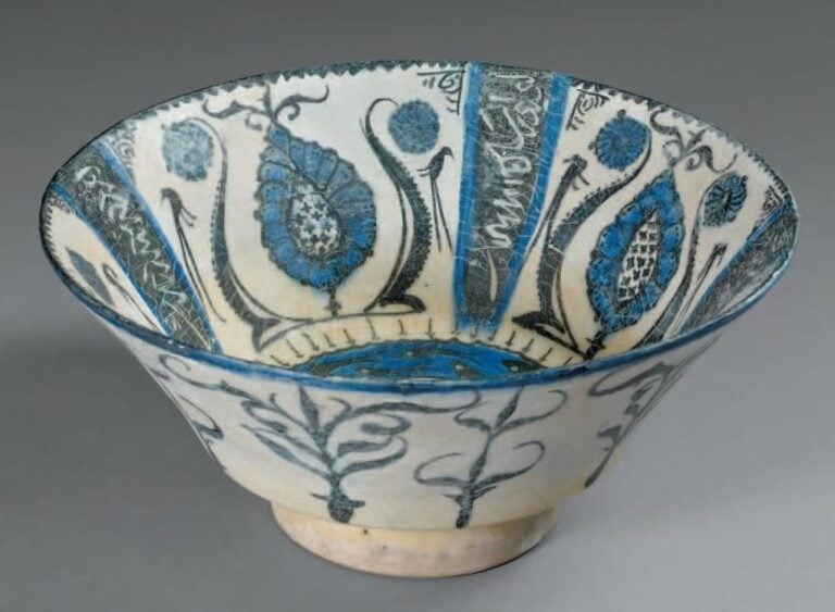 Coupe à décor rayonnant Céramique siliceuse à décor peint en bleu et noir sous glaçure transparente Cette coupe présente un décor très fin en bleu et noir sur fond blanc composé d'un médaillon central orné d'une palmette entourée de poissons et d'un bandeau où alternent un motif floral et des cartouches calligraphiques, l'inscription ici non déchiffrée correspond probablement à un extrait du Shahnamah de Firdousi, comme sur d'autres céramiques de ce typ