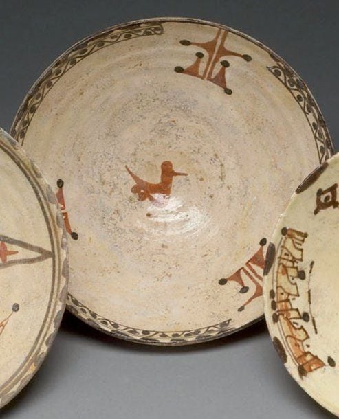 COUPE AUX INSCRIPTIONS COUFIQUES Coupe hémisphérique, décorée en rouge et brun sur engobe blanc d'un oiseau au centre et de trois inscriptions coufiques en pendentifs, séparées par trois cartouches de rinceaux ponctué
