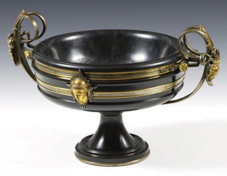 Coupe circulaire en marbre noir mouluré et bronze ciselé et doré; les anses curvilignes rattachées à des fleurettes prennent naissance de palmettes stylisées centrées de mascarons coiffés de serpents; la panse ornée d'un double bandeau rythmé de masques féminins se détachant de motifs stylisés; piédouche évasé à frises moulurée