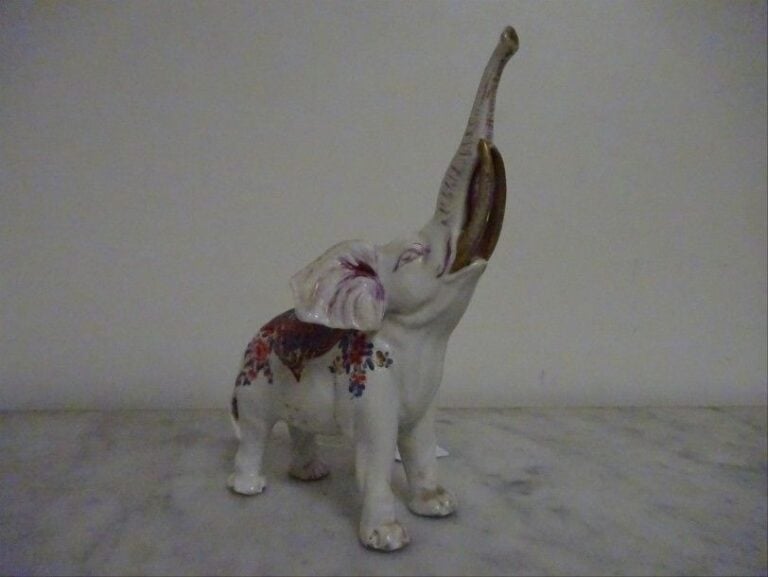ELEPHANT BLANC BARRISSANT EN PORCELAINE D'ARITA A DECOR KAKIEMON Japon, époque XIXe siècle, Meiji Représenté debout sur ses quatre pattes, la trompe relevée, la musculature nerveuse bien rendue dans un traitement naturaliste, défenses dorées ; porcelaine moulée rehaussée d'un décor émaillé de type Kakiemon, rinceaux fleuris et feuillagés de lotus sur le dos inscrits dans un médaillon quadrilobe flanqué de chrysanthèmes et de fleurs de prunus ; cavités sous les sabots laissant à penser que l'animal que nous présentons aurait pu coiffer une pendule ; l'éléphant blanc est un symbole d'espérance dans les croyances japonaises ? il est également partie prenante de l'iconographie bouddhiste en tant que monture du bodhisattva Fugen ; il s'agit là d'une iconographie typique de la production Kakiemon d'Arita : ainsi, dès la fin du XVIIe siècle, entre dans les collections d'Auguste Le Fort une sculpture de même type aujourd'hui conservée au Musée de Dresd