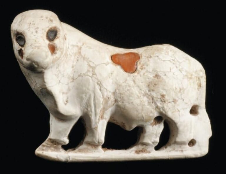 FIGURINE DE BOVIDÉ Figurine en calcite blanche, à dos plat, représentant un bovidé passant sur une barre, tête tournée de côté, le corps et la tête incrustés de pierre