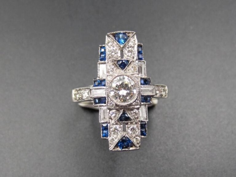 Fine BAGUE rectangulaire en or gris (750 millièmes) serti de diamants taille brillant et baguettes et saphirs calibrés, à motifs géométriques             Long        motif : 2,1 cm        Doigt : 52        Poids brut : 7 g