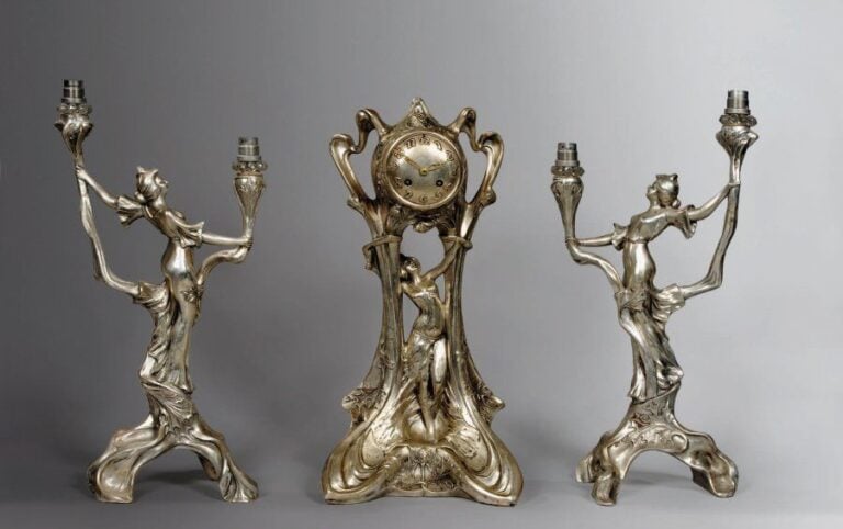 Garniture de cheminée en régule argenté (composition) trois pièces à décor de femmes et de chardons en haut reliefs entourés d'arborescence ondulées se composant d'une pendule (sans garantie de marche) et d'une paire de flambeaux à deux bras de lumière tenus par une femme debou