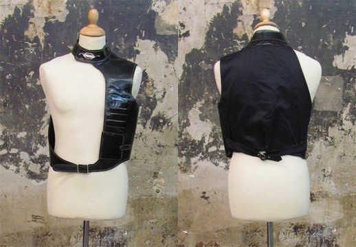 GILET sans manches asymétrique pour Homme en simili cuir noir orné de surpiqûres blanches et satin à la couleur, petit col droit se pressionnant, parementure gauche agrémentée de poches superposées puis d'un rabat contenant un portefeuille, taille s'attachant à l'aide d'une boucle, pattes de serrage au dos (griffe noire, graphisme vert) (T50)