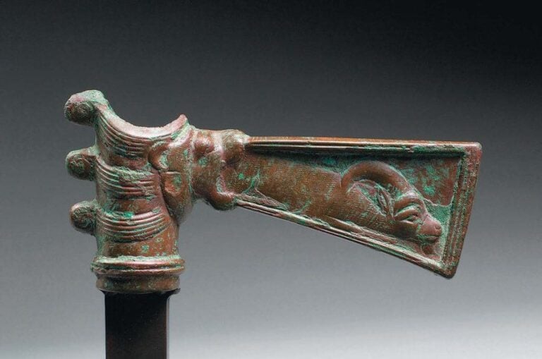 HACHE AUX ANIMAUX Hache à douille cylindrique dont les nervures se prolongent sur le talon par trois volutes et ornée à l'opposé d'une tête de lionne dont la gueule béante crache une lame trapézoïde ornée en léger relief sur les deux faces d'un bouquetin couch