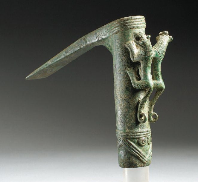 HERMINETTE AUX LIONS Superbe herminette à lame aplatie et douille tubulaire moulurée vers le bas, ornée de rainures en « dents de scie » et de cercles concentriques en relief et flanquée sur le talon d'un couple de lions debout, tête tournée, leur crinière figurée par de fins chevrons gravé