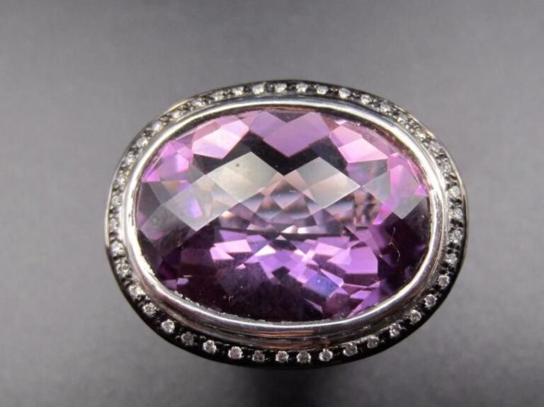 Importante BAGUE en or gris et or noirci (750 millièmes) ajouré à motifs de volutes surmontée d’une améthyste ovale facettée entourée de petits diamants taille brillant             Doigt : 53-54        Poids brut : 13,8 g