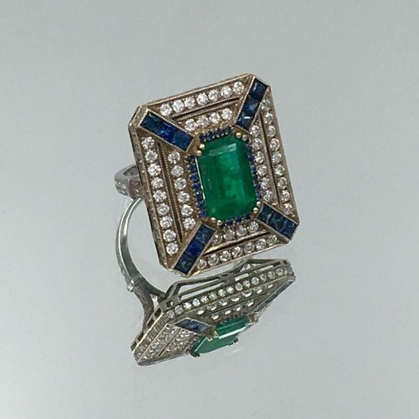 Importante BAGUE rectangulaire en or gris (750 millièmes) finement ajouré, centrée d'une émeraude rectangulaire taillée à degrés, entourée de diamants taille brillant, de saphirs calibrés rond