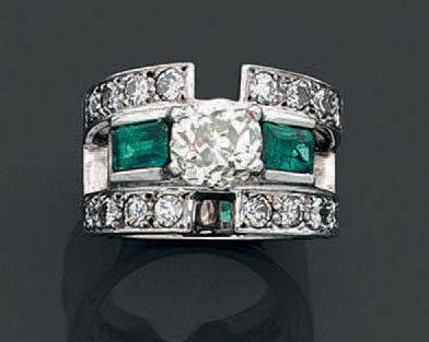 Large BAGUE en or gris (750 millièmes) ajouré serti au centre d'un diamant taille ancienne de forme coussin épaulé de deux émeraudes rectangulaires taillées à degrés, soulignées de diamants taille brillant de part et d'autr