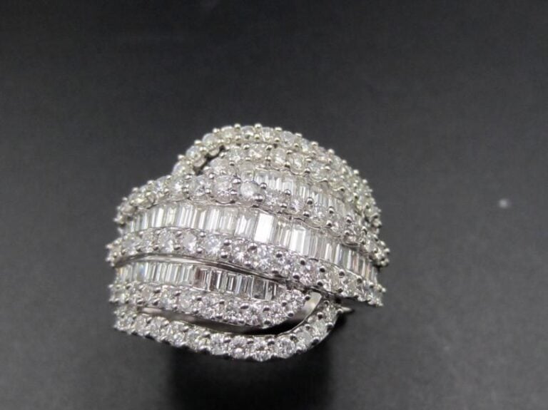 Large BAGUE « volute »  en or gris (750 millièmes) ajouré serti d’alignements de diamants baguettes alternés de diamants taille brillant              Doigt : 56         Poids brut : 9,9
