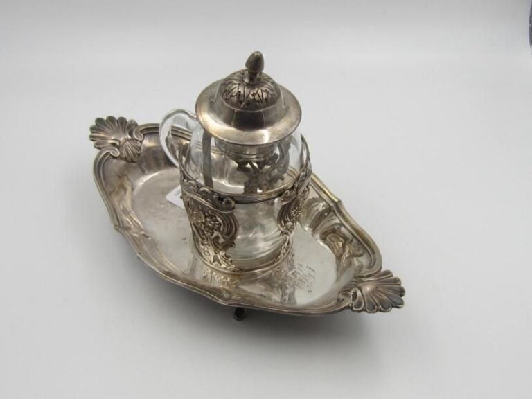 Moutardier en argent posant sur un plateau ovale aux bords chantournés terminés par un motif de coquille à chaque extrémité; le corps du moutardier ajouré de grappes de raisin dans des cartouches feuillagés; quatre petits pieds à enroulements; intérieur en verre blanc; Monogramme «P» sur le plateau; bouchon à décor feuillagé, frétel en graine (petite restauration)