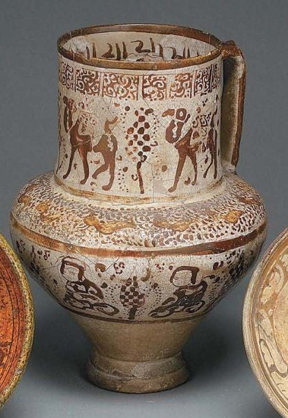 PICHET À DÉCOR FIGURATIF Pichet à panse tronconique, col cylindrique et anse verticale, décoré en lustre métallique brun sur fond blanc de sept personnages assis parmi des cyprès le long de la panse, d'une frise d'oiseaux sur l'épaule, d'un personnage et de quatre chameaux passant autour du co