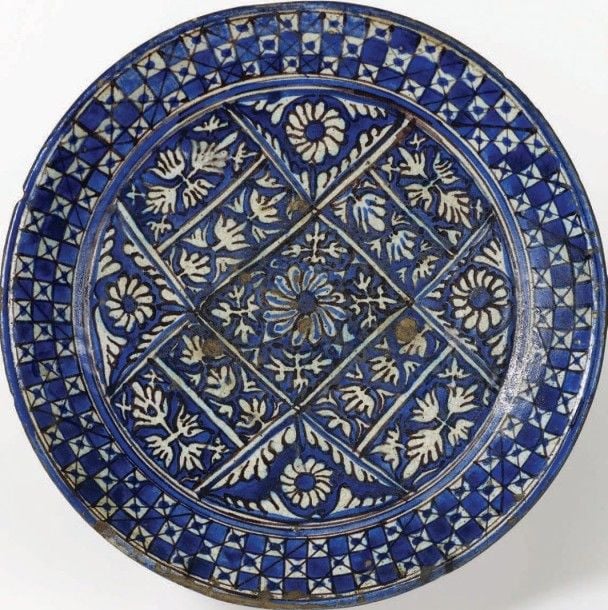 Plat à décor floral Terre cuite à décor peint en bleu, blanc et noir Asie Centrale, Probablement Boukhara, XVIIIe siècle Diamètre: 32,5 cm Il est décoré de fleurs et de feuilles dans une structure losangée, le marli est orné d'une mosaïque de carré
