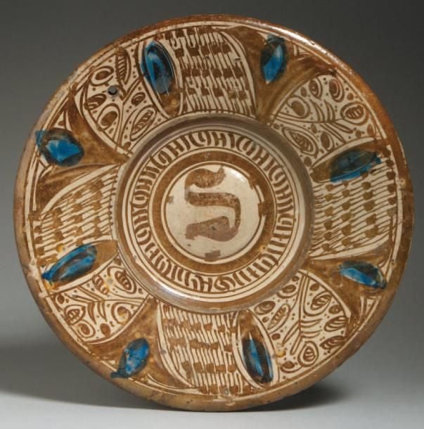 PLAT LUSTRÉ Plat décoré en lustre métallique brun sur fond blanc d'un bandeau pseudo-épigraphique autour d'un ombilic et de bourgeons bleus en léger relief entourés de motifs floraux et « notes » noire