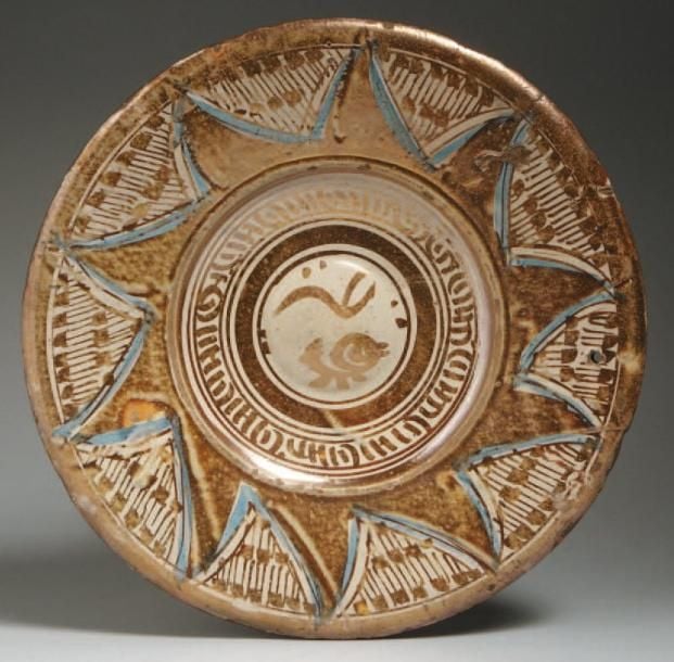 PLAT LUSTRÉ Plat décoré en lustre métallique brun sur fond blanc d'un bandeau pseudo-épigraphique autour d'un ombilic et de triangles moulés cernés de bleu, formant une rosace rayonnante sur un champ de « notes » noire