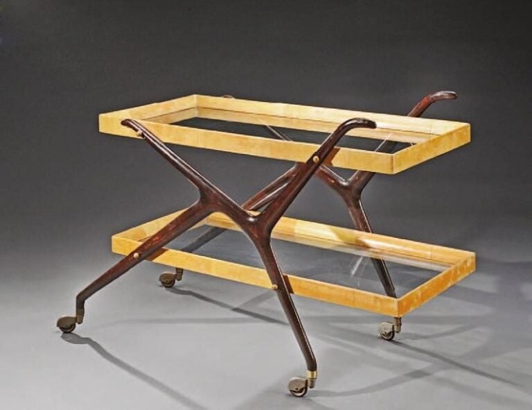 Table desserte à deux plateaux rectangulaires en verre dans des encadrements de bois gainés de parchemin, les montants en bois sombre dessinant un «K» sont terminés par des roulette