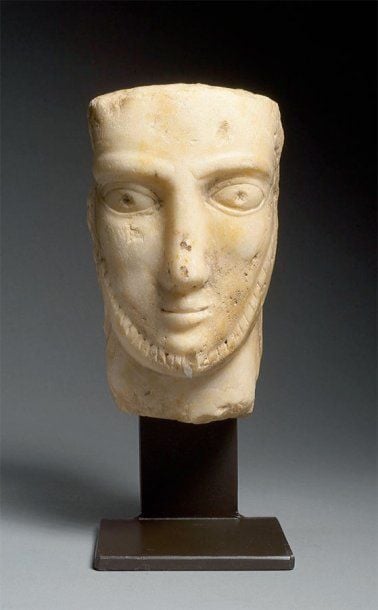 TÊTE D'HOMME Tête en marbre, au visage triangulaire, sourcils en relief, yeux en amande à l'iris ponctué, nez droit, bouche petite et barbe en collier gravée de strie