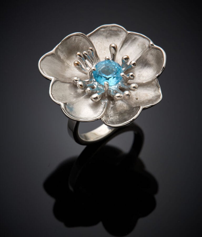 BAGUE d'artiste "fleur" en or gris (750‰) satiné, centré d'une topaze bleue ron…