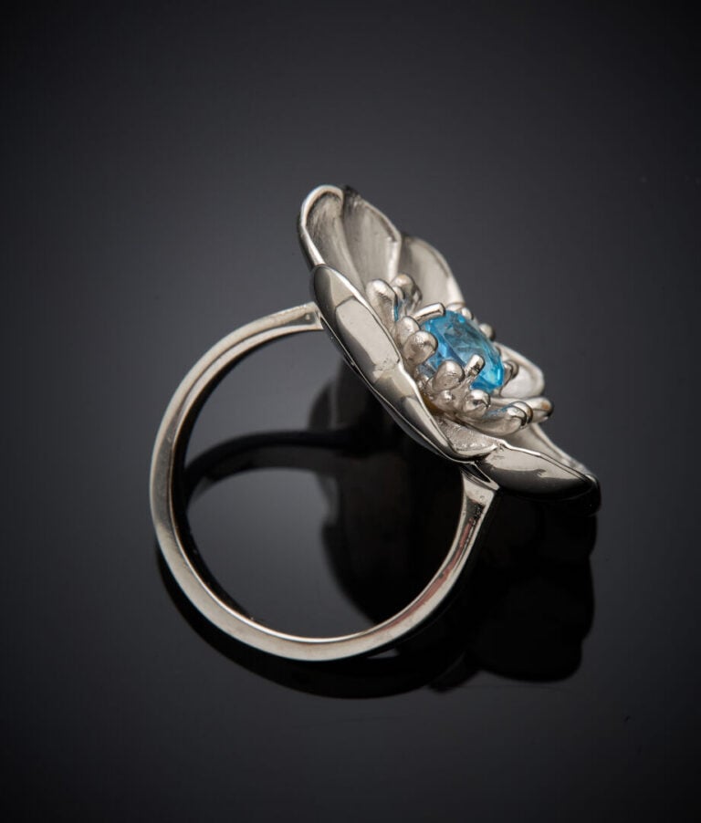 BAGUE d'artiste "fleur" en or gris (750‰) satiné, centré d'une topaze bleue ron…