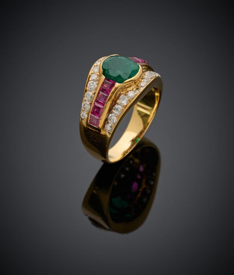 Large BAGUE en or jaune (750‰) serti d'une émeraude ovale, soulignée de rubis c…